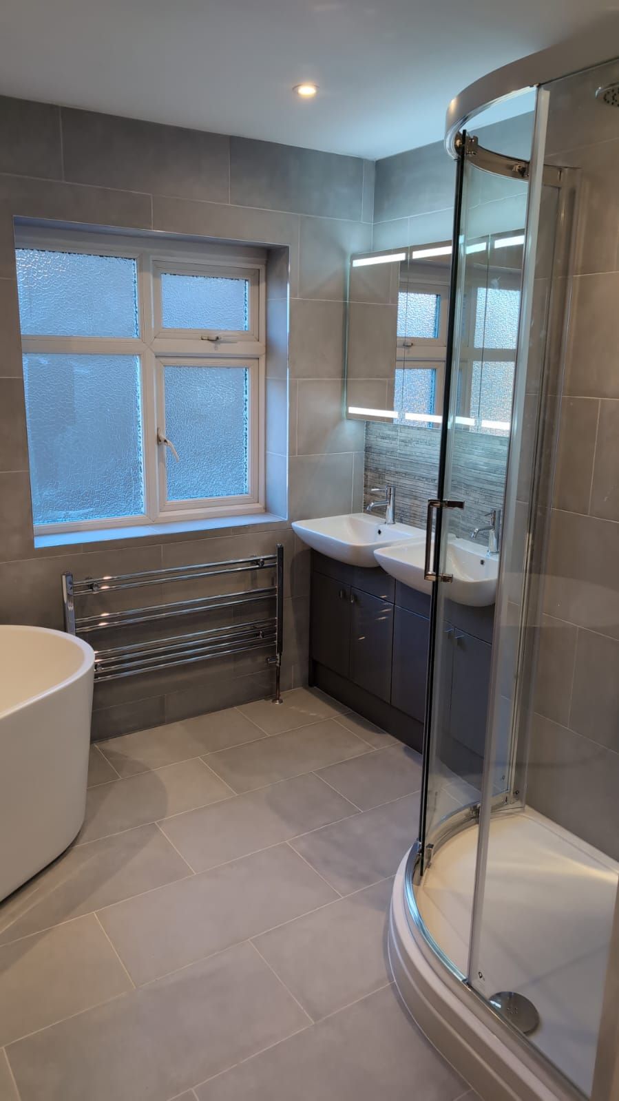 Finished bathroom featuring a curved shower enclosure, double basin vanity, freestanding bath and grey floor and wall tiles