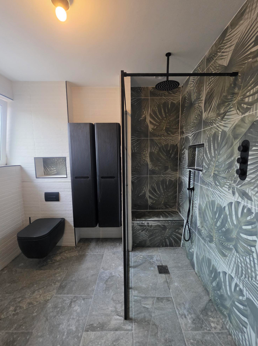 A bold botanical wet room featuring Bonito Jungle leaf-print tiles, an Amien matt black grid shower screen, and a Micheli wall-hung toilet alongside Soar ribbed black storage cabinets and Dosimo Musgo stone floor tiles.