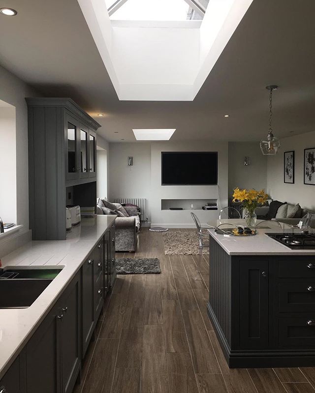 A vast open-plan living area featuring dark grey shaker-style kitchen cabinetry and a matching island, unified by rich brown wood-effect floor tiles. The space is flooded with natural light from large skylights and transitions into a cosy lounge area.