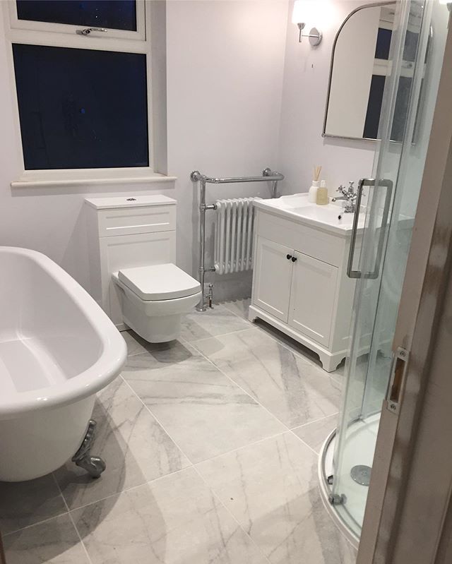 A classic white roll top bath with chrome feet sits on large marble-effect floor tiles, next to a matching white traditional vanity unit and a period-style chrome and white towel radiator.