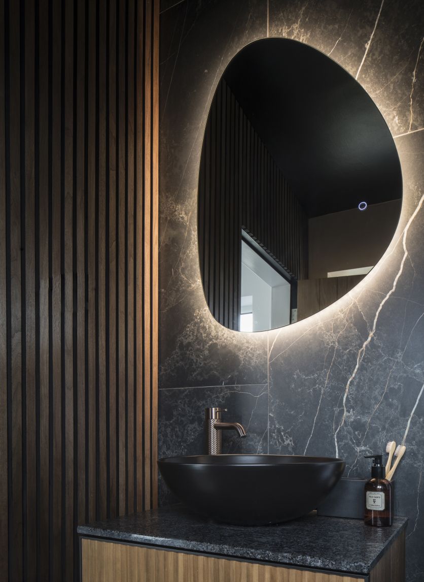 A sophisticated ensuite featuring a matt black countertop washbowl on a dark stone surface, set against a backdrop of dramatic black marble wall tiles and vertical wood slat panelling.