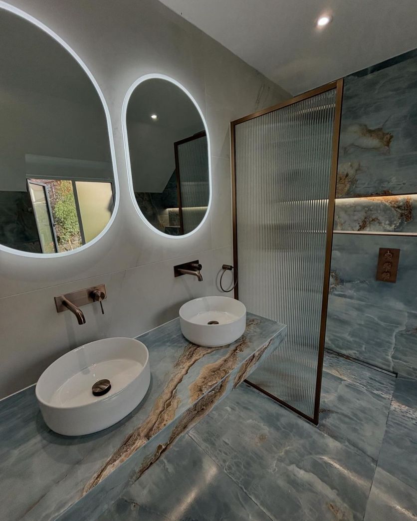 A high-end bathroom featuring blue onyx-effect porcelain tiles, a double vanity with Damon white countertop basins, Moray satin bronze knurled taps, and an Amien fluted glass shower screen.