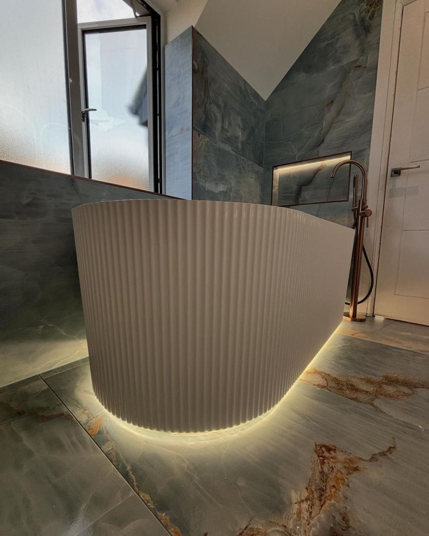 A luxurious industrial-style bathroom featuring the Olympia white ribbed freestanding bath with glowing under-bath LED lighting, set against dramatic blue marble-effect wall and floor tiles with a Moray satin bronze floor-standing tap.