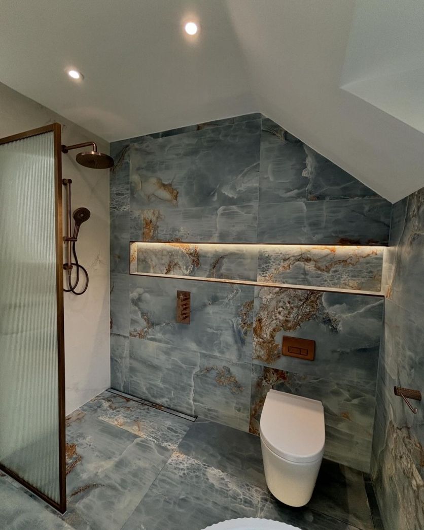 A high-end style wet room featuring blue marble-effect tiles, an Amien brushed bronze framed fluted glass screen, and Moray satin bronze shower fittings alongside a Froxfield wall-hung toilet.