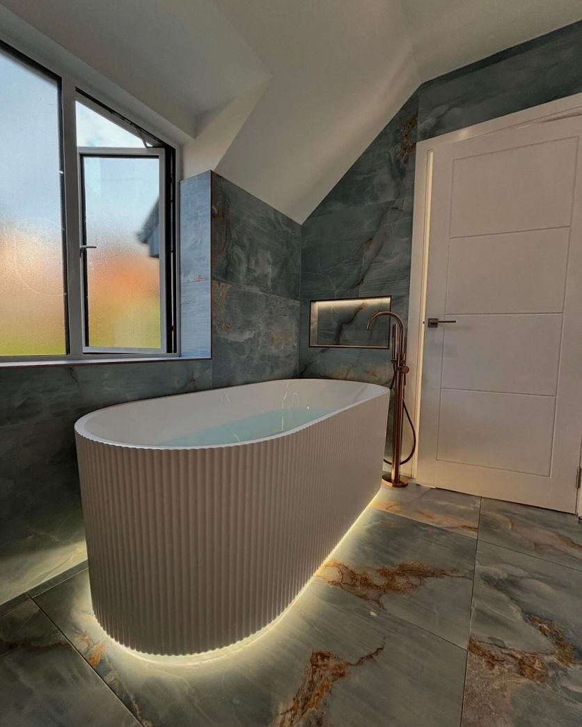 A luxurious industrial-style bathroom featuring the Olympia white ribbed freestanding bath with glowing under-bath LED lighting, set against dramatic blue marble-effect wall and floor tiles with a Moray satin bronze floor-standing tap.