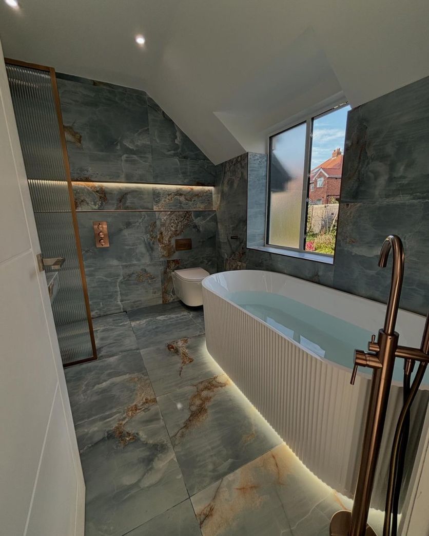 A stunning industrial-inspired bathroom featuring blue marble-effect tiles on walls and floor, an Amien brushed bronze framed fluted glass shower screen, and an Olympia white ribbed freestanding bath with a Moray satin bronze floor-standing tap.