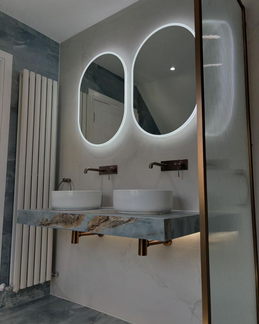 A high-end bathroom featuring a floating blue marble-effect vanity with twin Damon white countertop basins, Moray satin bronze wall-mounted taps, and Halle oval illuminated mirrors against a white vertical radiator and fluted glass screen.