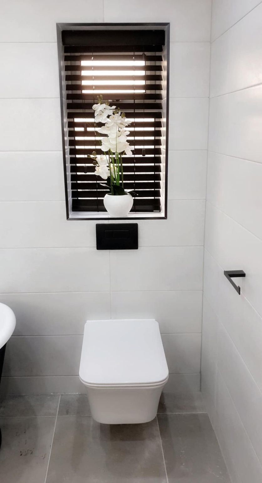 Wall hung white toilet with matte black flush plate and accessories, framed window with black blinds and concrete-effect grey floor tiles.
