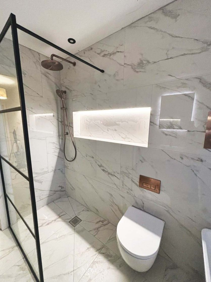 A contemporary wet room featuring a Stockholm Odlina white marble-effect gloss porcelain tiled wall and floor, accented by an Amien matt black framed shower screen and a Moray satin bronze concealed shower set.