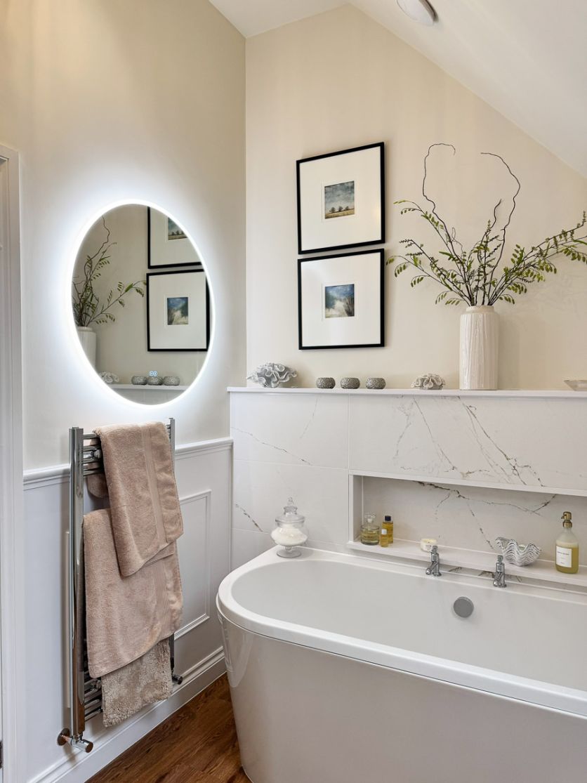A bright, neutral bathroom featuring a large Felix round backlit LED mirror, a Neptune chrome heated towel rail, and Grosvenor chrome bath pillar taps set against Dune marble-effect wall tiles.