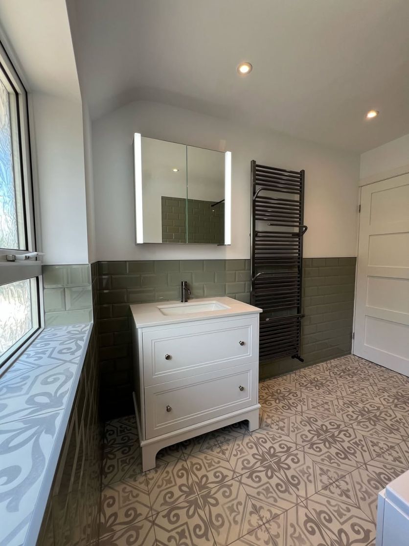 A traditional Ferris white floor-standing vanity unit with a quartz worktop and Moray gunmetal knurled tap, set against sage green metro wall tiles and Xabia silver patterned floor tiles.