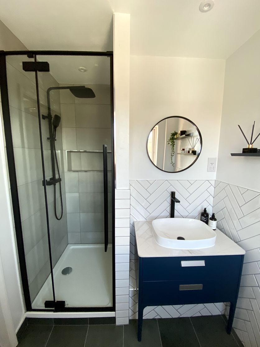 A modern bathroom featuring a black framed shower enclosure with a matt black rainfall shower, alongside a navy blue vanity unit with a white countertop basin and a black tall mixer tap, set against white herringbone wall tiles.