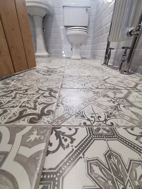 Intricate grey and white patterned floor tiles leading towards a traditional white pedestal basin and a classic close-coupled toilet, framed by white gloss metro wall tiles and a chrome heated towel rail.