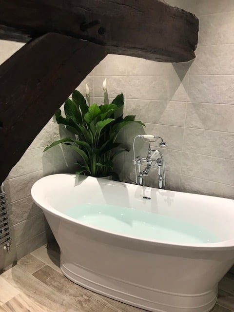 Freestanding white bath beneath rustic wooden beam with chrome standpipe tap, textured wall tiles and leafy plant.