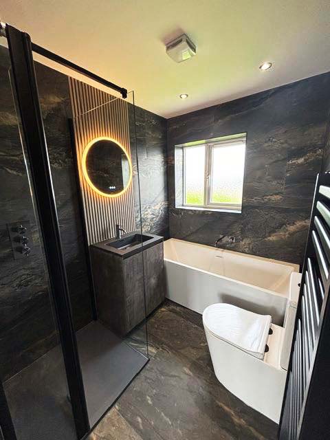 A dramatic modern bathroom featuring Duomo dark grey marble-effect porcelain wall tiles and an Ottoman slatted wood-effect feature wall, accented by a Moray gunmetal shower set and an Odion metallic floor-standing vanity with a black basin.