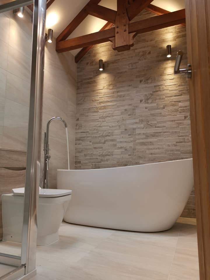 A modern white freestanding slipper bath with a curved chrome floor-standing tap, set against a textured beige split-face stone feature wall under original wooden ceiling beams with contemporary cylinder spotlights.