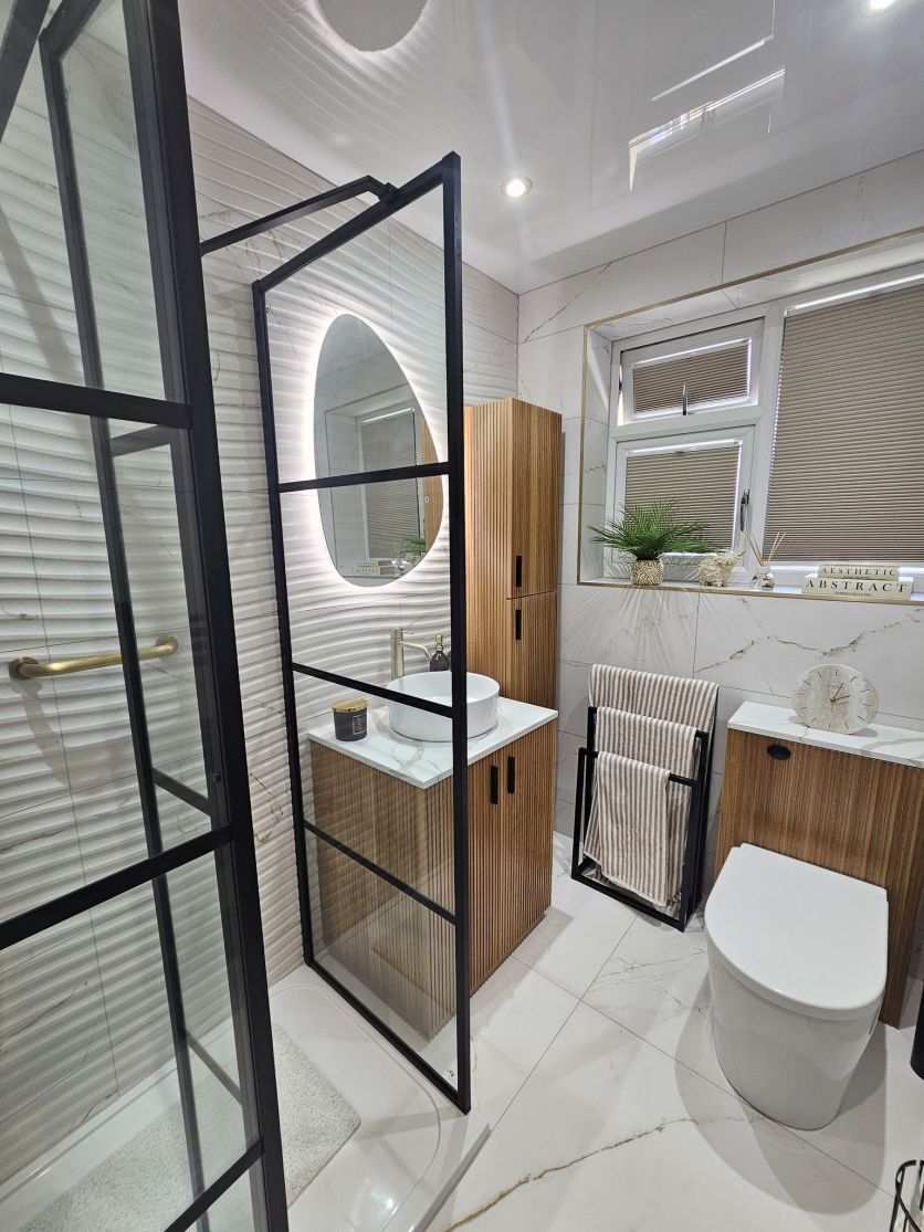 A designer bathroom display featuring a Revel walnut fluted vanity, an Amien black-framed glass shower screen, and Dune 3D textured wall tiles accented by an Ivy pebble shaped LED mirror.