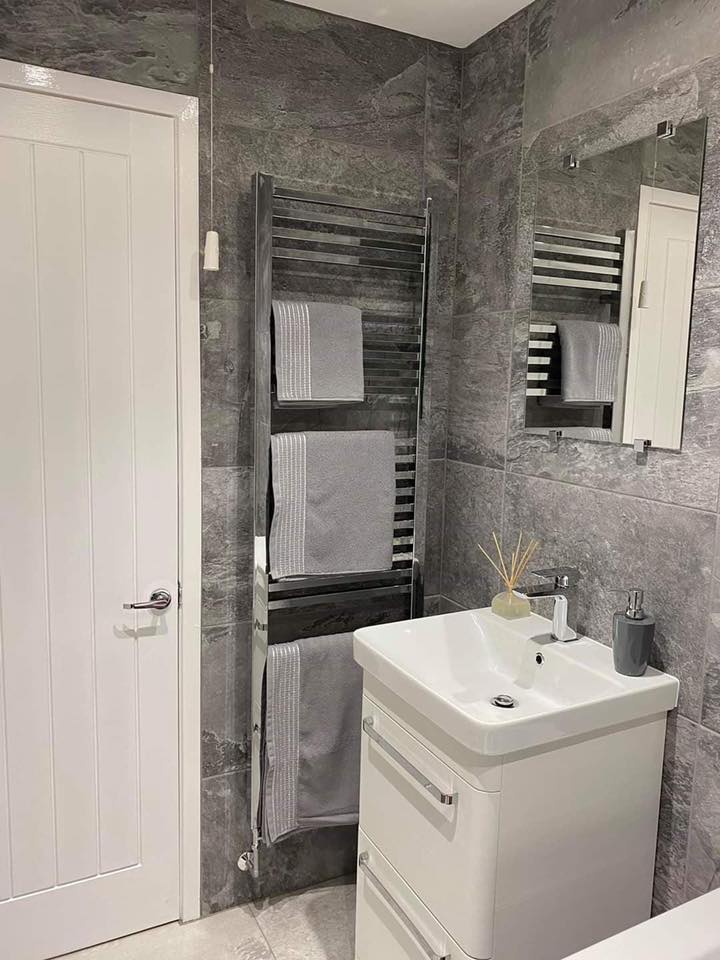 A floor-standing white gloss vanity unit with chrome handles and a square basin, paired with a tall chrome heated towel rail against grey slate-effect tiles.
