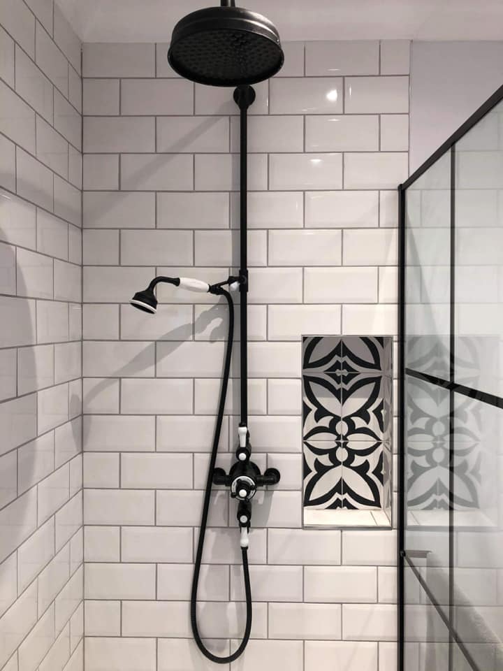 A sophisticated traditional shower featuring an exposed matt black rainfall shower kit with white ceramic handles. The space is finished with white metro wall tiles and a decorative recessed niche showcasing black and white patterned tiles.