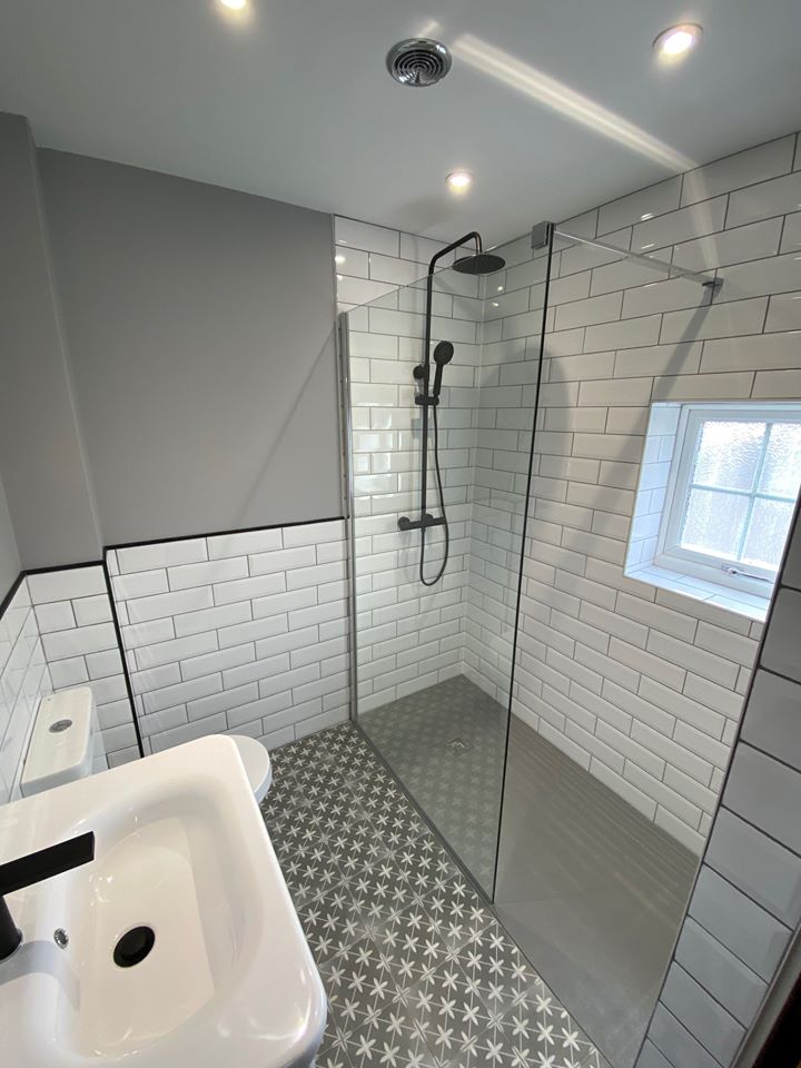 A contemporary walk-in wet room featuring white metro wall tiles with contrasting dark grout, a sleek matte black rainfall shower head, and grey patterned Victorian-style floor tiles.