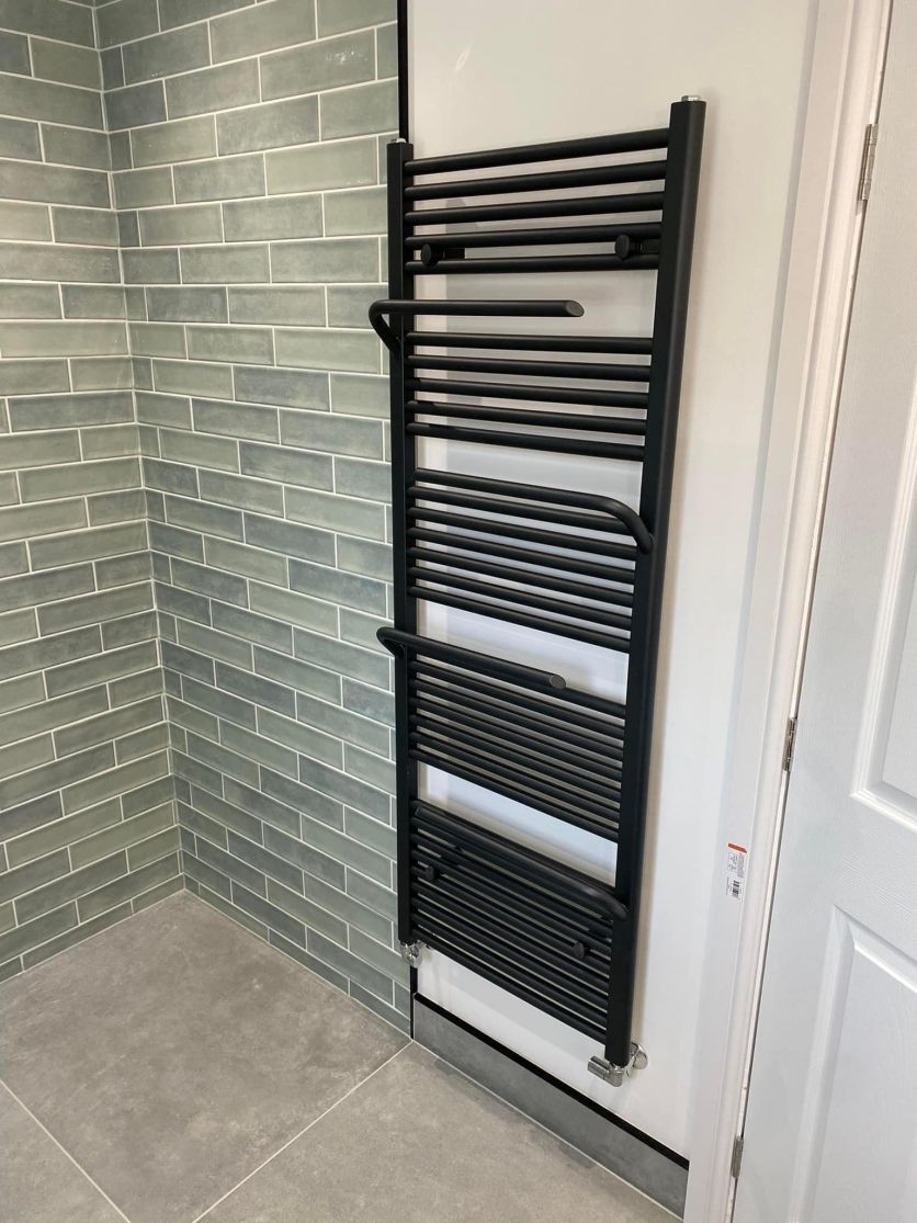A contemporary tall matt black heated towel radiator with integrated curved hanging rails, mounted against a crisp white wall adjacent to soft sage green metro wall tiles