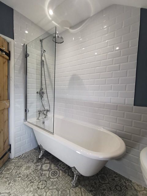 An elegant white freestanding roll top bath with chrome claw feet, featuring a glass shower screen and traditional chrome mixer shower, set against white bevelled metro wall tiles and grey patterned Victorian-style flooring.