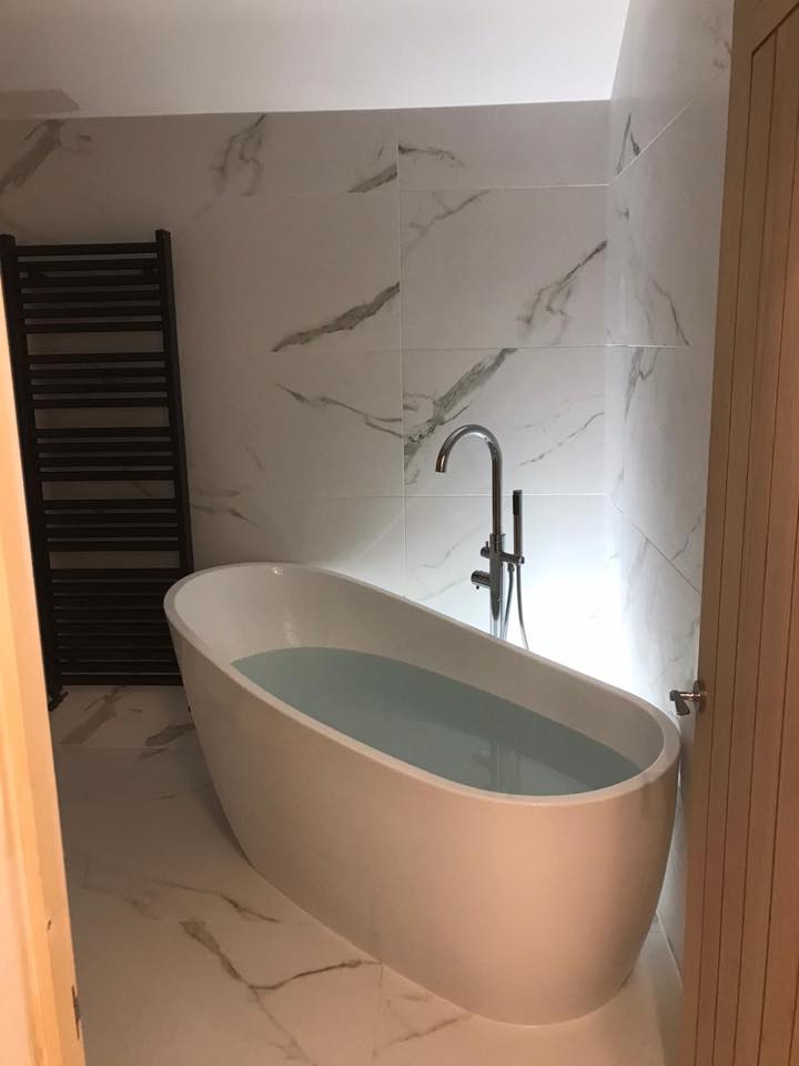 An elegant white oval freestanding bath with a floor-standing chrome swan-neck mixer tap, set against large-format white marble-effect wall and floor tiles with grey veining, featuring a contrast black heated towel rail.