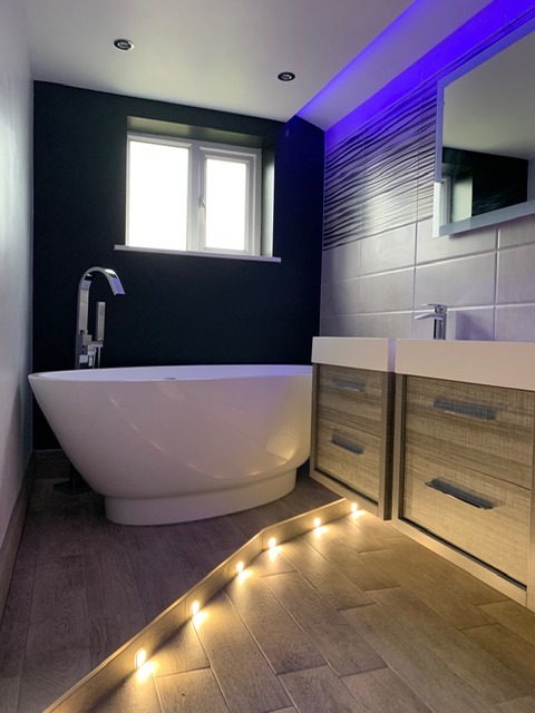 An elegant white oval freestanding bath with a chrome floor-standing mixer tap, situated next to grey wood-textured floating vanities. The space is illuminated by purple ceiling LEDs and warm white recessed floor plinth lights for a high-end spa feel.