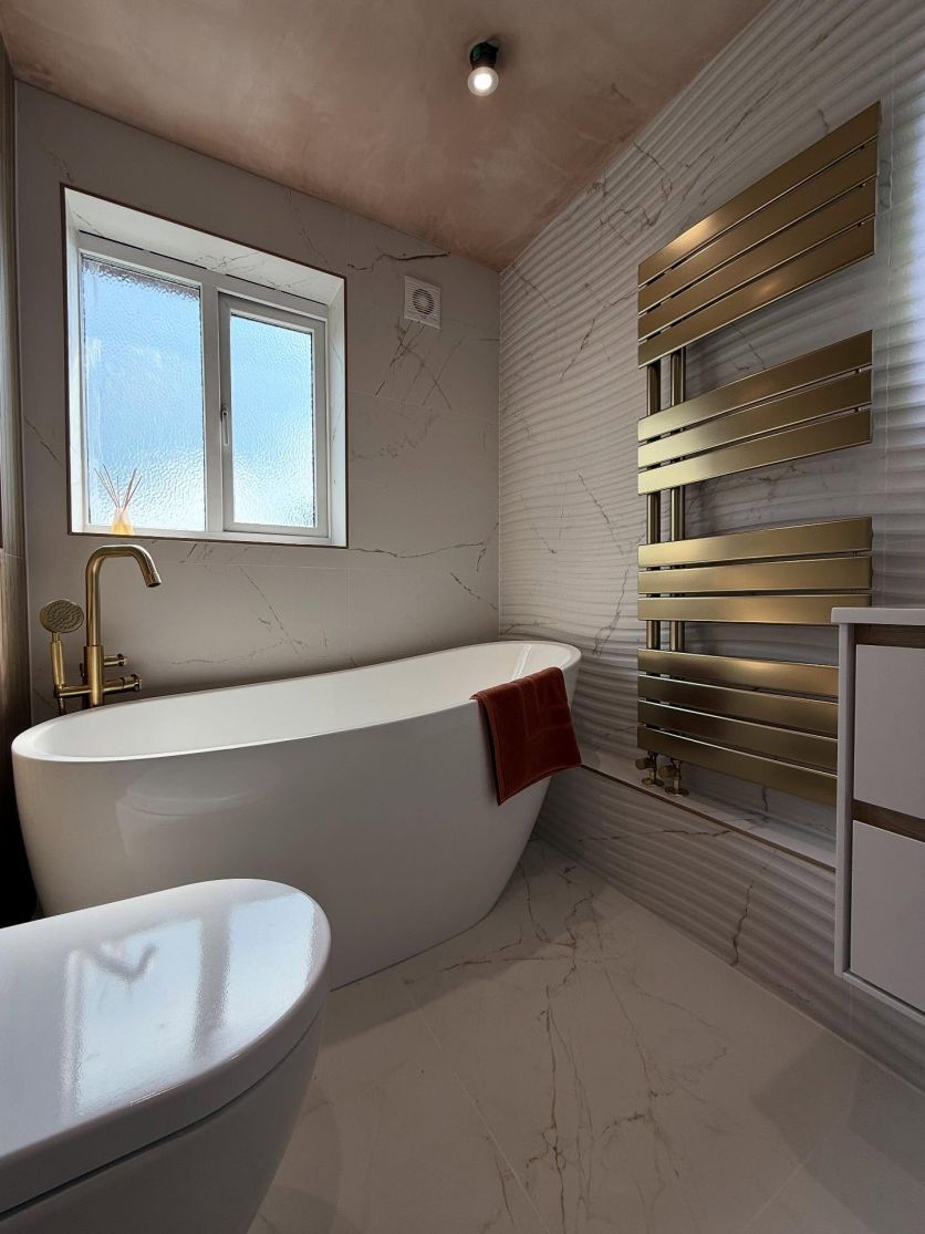 A luxury boutique bathroom featuring an Isleworth freestanding slipper bath with an Urban brushed gold mixer tap, set against Dune marble 4D textured wall tiles and a Tarvos brushed gold heated towel rail.