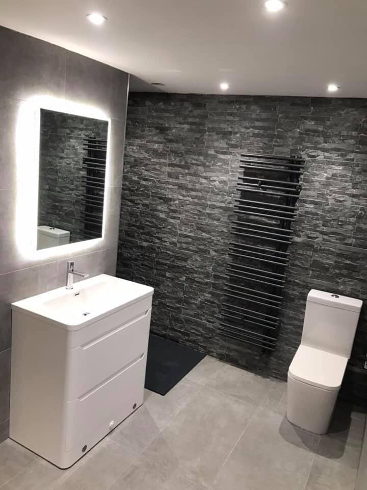 A modern monochromatic bathroom featuring a textured slate split-face feature wall, an anthracite designer heated towel rail, and a floor-standing white gloss vanity unit with a chrome mixer tap, topped by a large rectangular LED illuminated mirror.