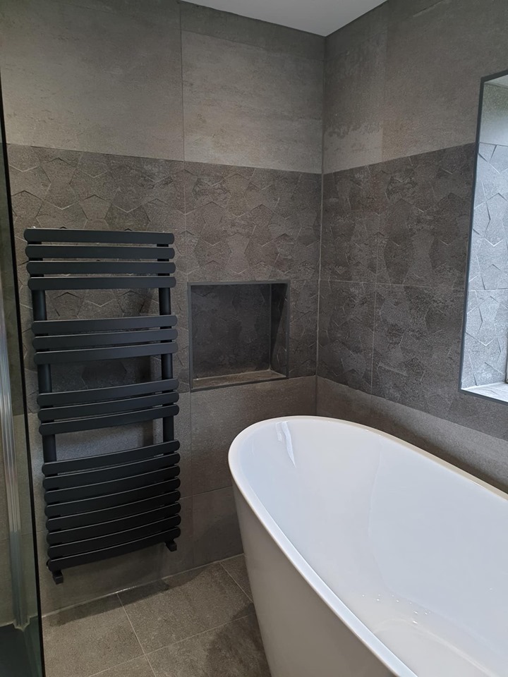 Modern grey bathroom with textured feature tiles, built-in wall niche, anthracite towel rail and a sleek freestanding bath.