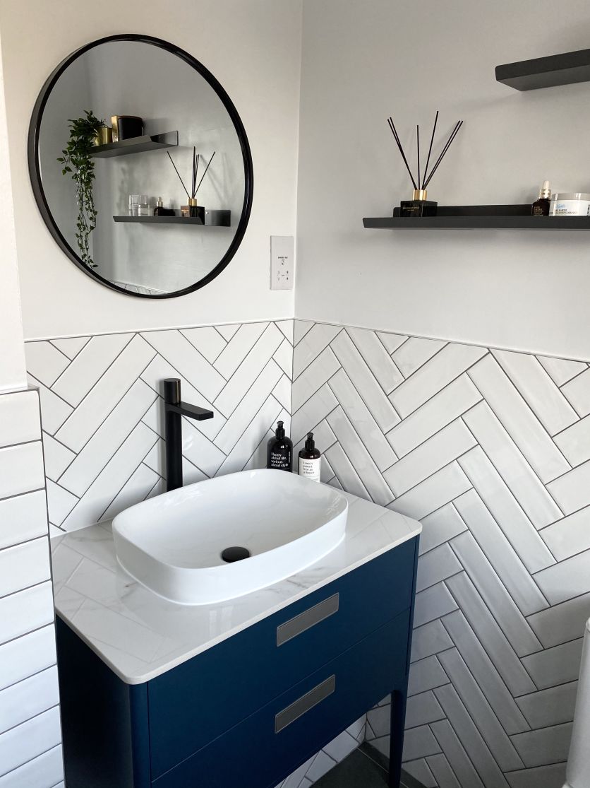Modern bathroom with white herringbone tiles, a petrol blue vanity, matt black tap and a round black-framed mirror.