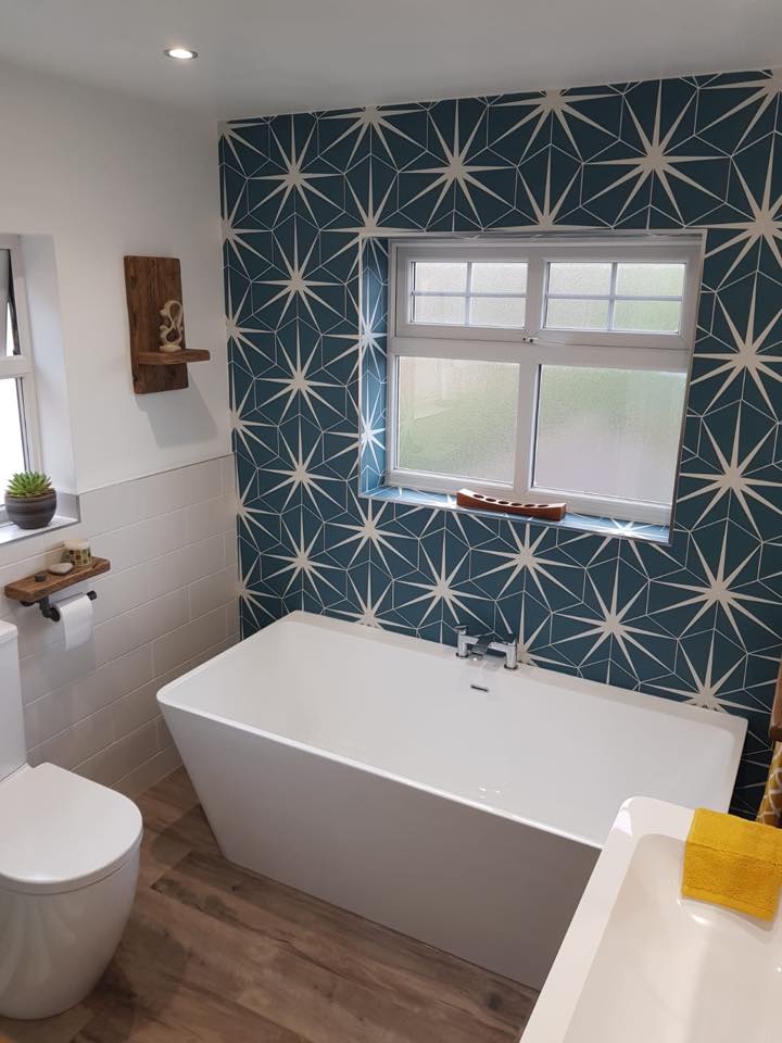 A modern white freestanding bath set against a striking blue geometric star-patterned feature wall, complemented by warm wood-effect flooring and rustic timber shelving.