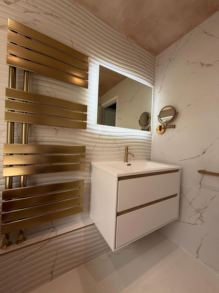 A luxury bathroom feature wall showcasing Dune Marble 4D decor matt ceramic tiles with sinuous textured curves, a Newbury wall-hung vanity in matt white and sawn oak, and a Tarvos brushed gold heated towel rail.