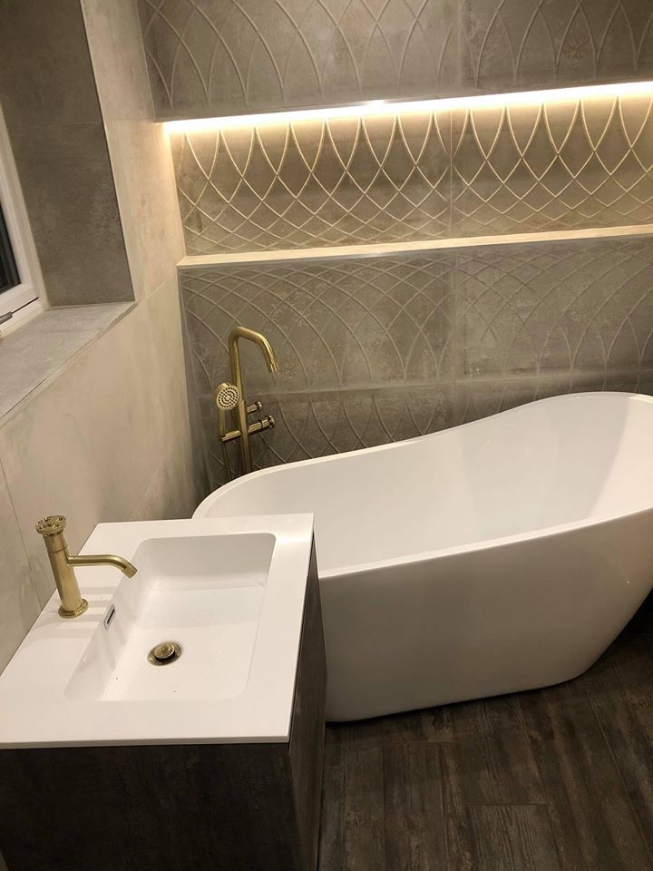 A modern white freestanding slipper bath paired with a floor-standing brushed gold bath mixer tap, set against decorative arched textured wall tiles with integrated LED strip lighting.