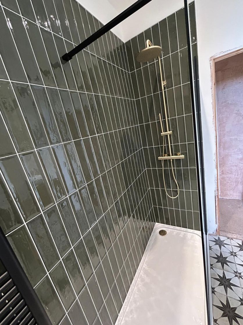 A modern walk-in shower featuring Navajo Oliva gloss olive green tiles in a vertical brick layout, paired with an Elora brushed gold thermostatic shower and Fired Star patterned floor tiles.
