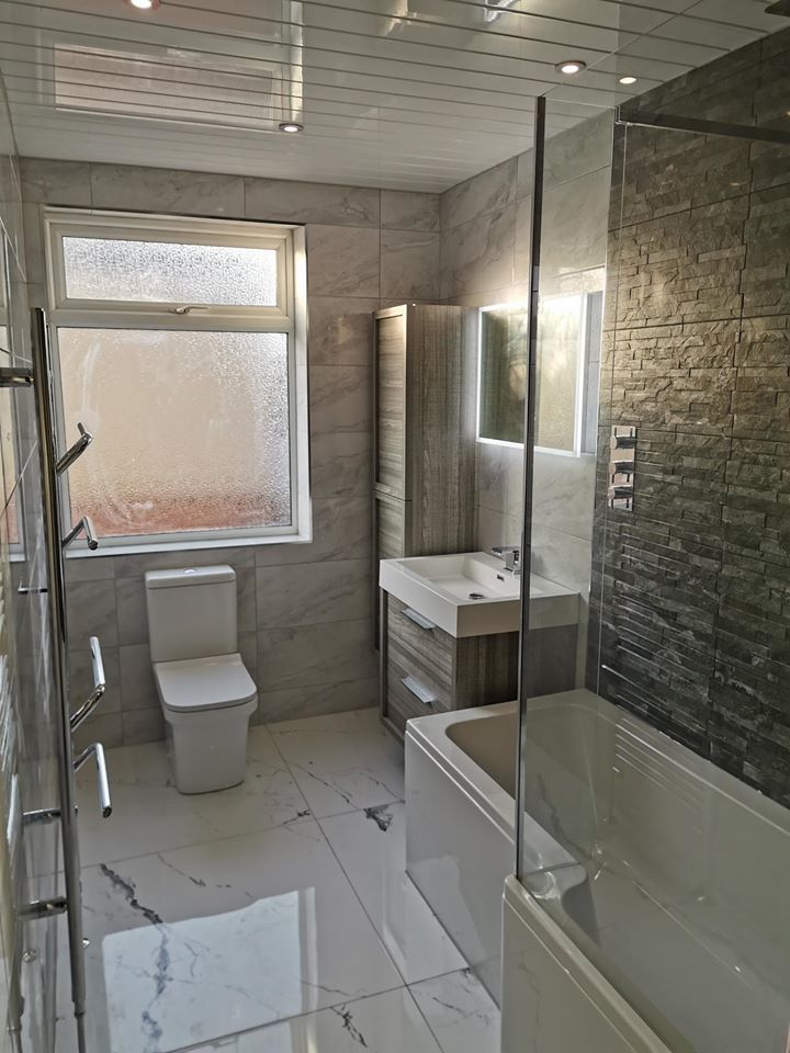 A complete modern bathroom featuring a grey wood-effect vanity unit and matching tall cabinet. The space includes an L-shaped shower bath with a slate-textured feature wall