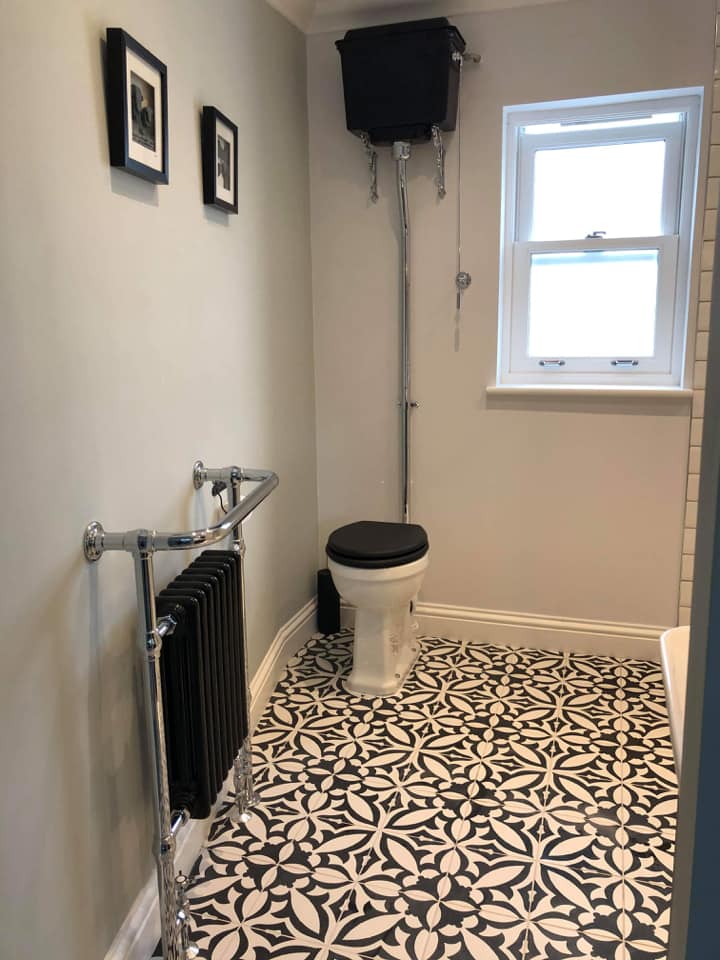 A classic heritage-style bathroom featuring a high-level toilet with a black cistern and matching black seat. The space is finished with striking monochrome patterned floor tiles and a traditional black column radiator with a chrome heated towel rail.