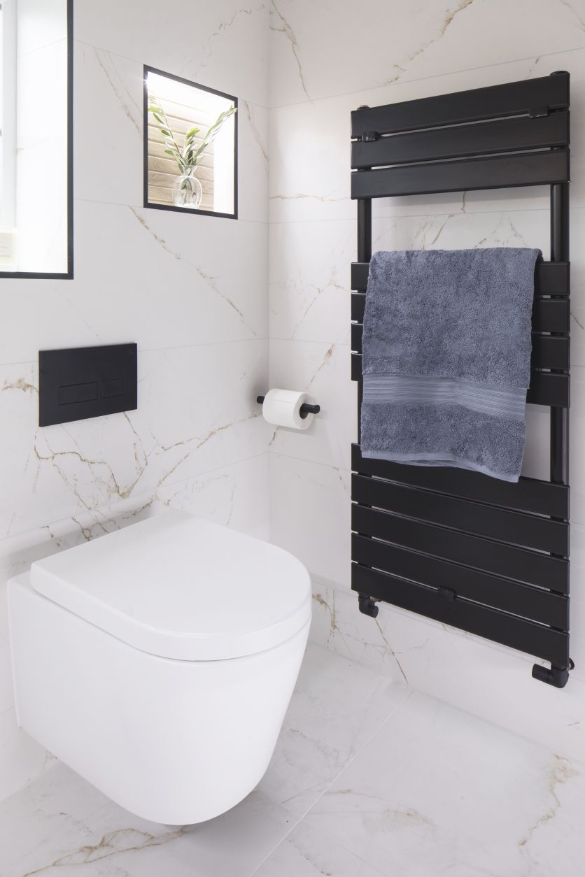 A minimalist wall-hung white toilet with a matt black dual flush plate, situated next to a designer black heated towel rail. The space is finished with large-format white marble wall tiles featuring delicate gold veining and a recessed shelf.