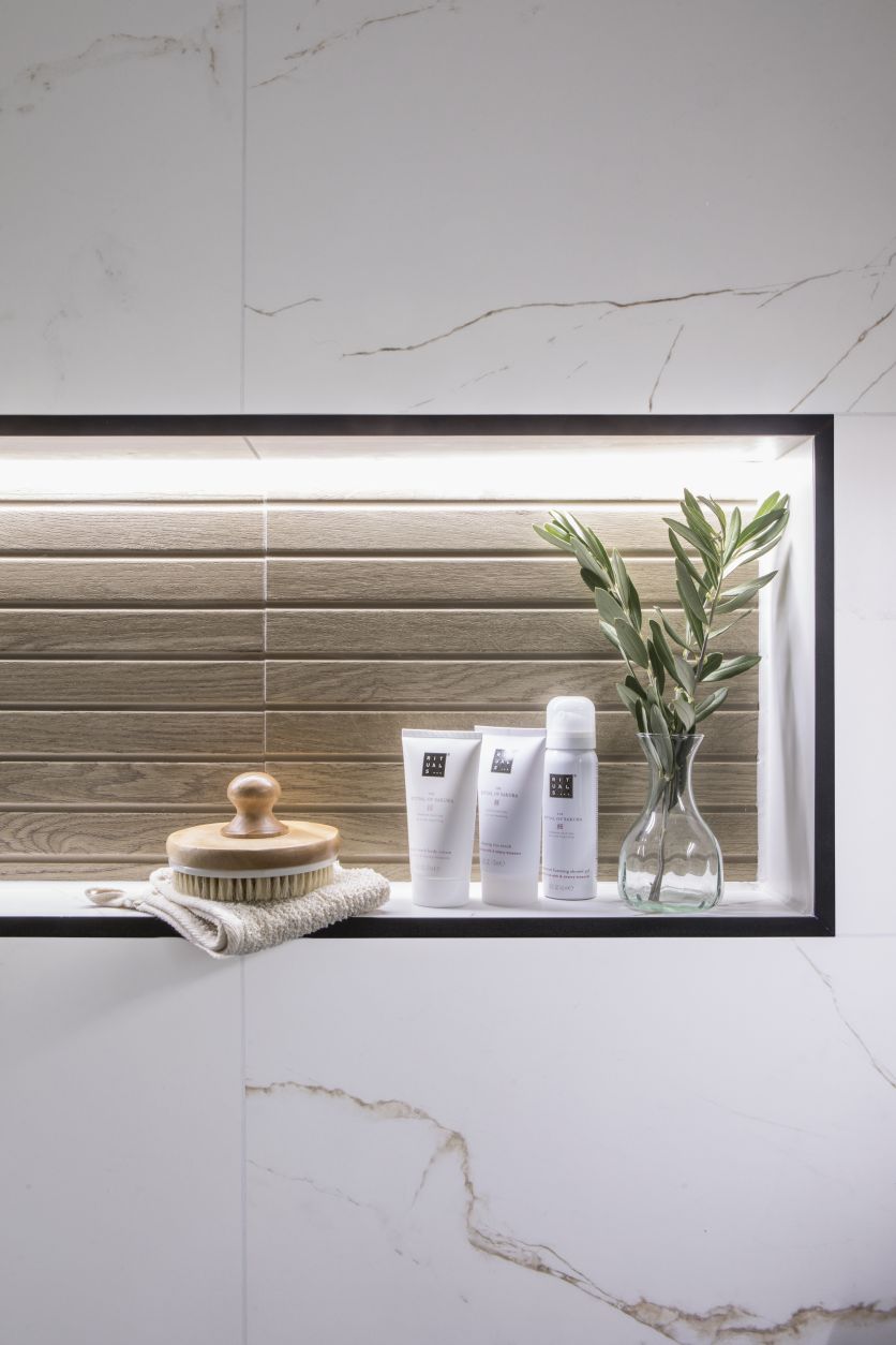 A close-up of an illuminated recessed bathroom niche featuring a black frame and textured oak wood-effect tiles, set into a wall of large-format white marble tiles with delicate gold veining.