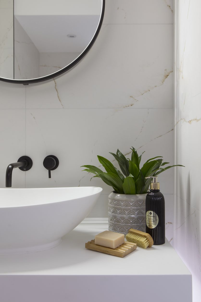 A sleek white countertop vessel basin paired with wall-mounted matt black mixer taps, set against large-format white marble wall tiles with delicate gold veining. A black-framed round mirror and lush green plant complete the minimalist look.