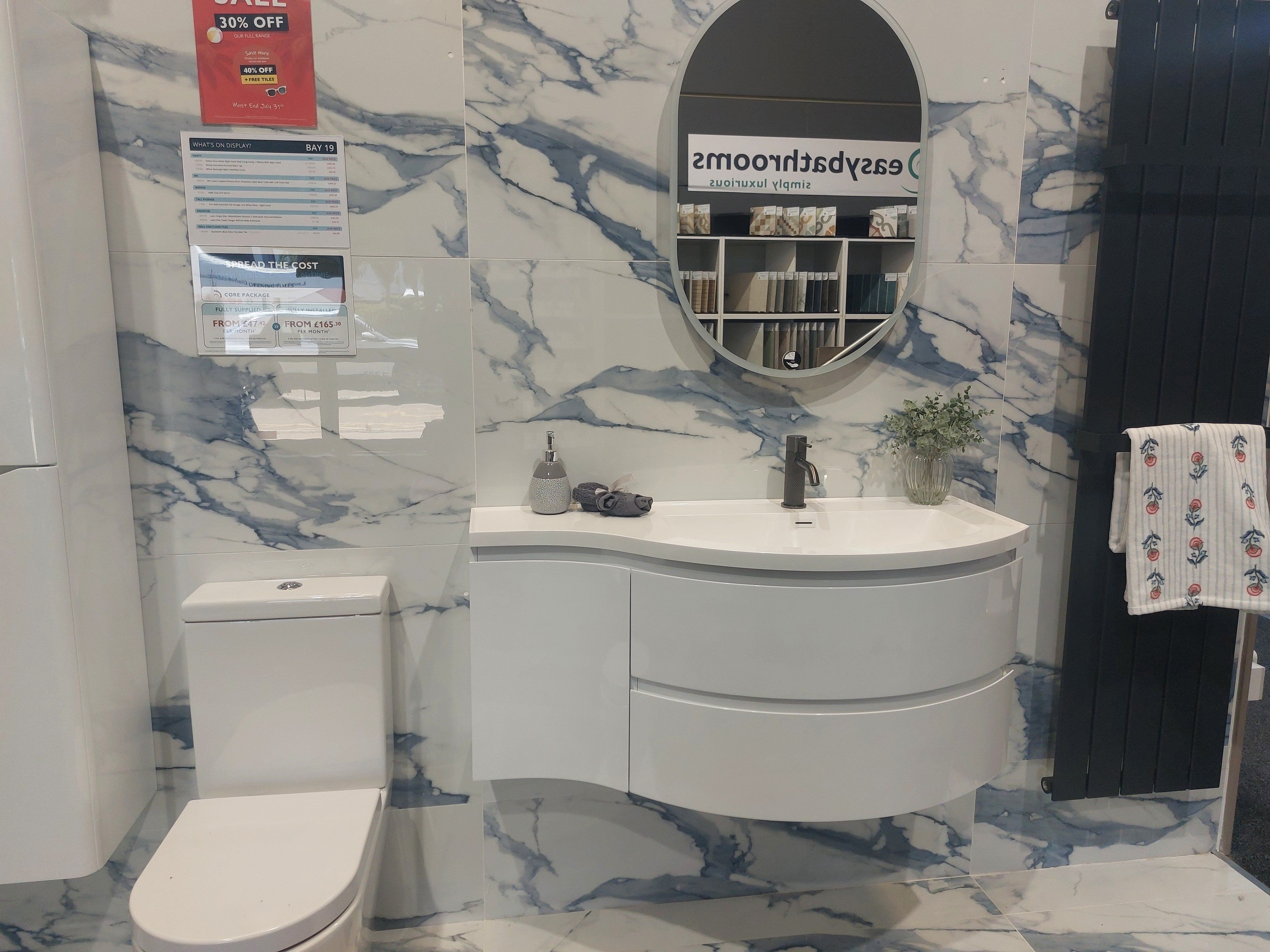Blue marble-effect bathroom display with a curved white vanity unit, round mirror, toilet and dark radiator at the Easy Bathrooms Huntingdon showroom.