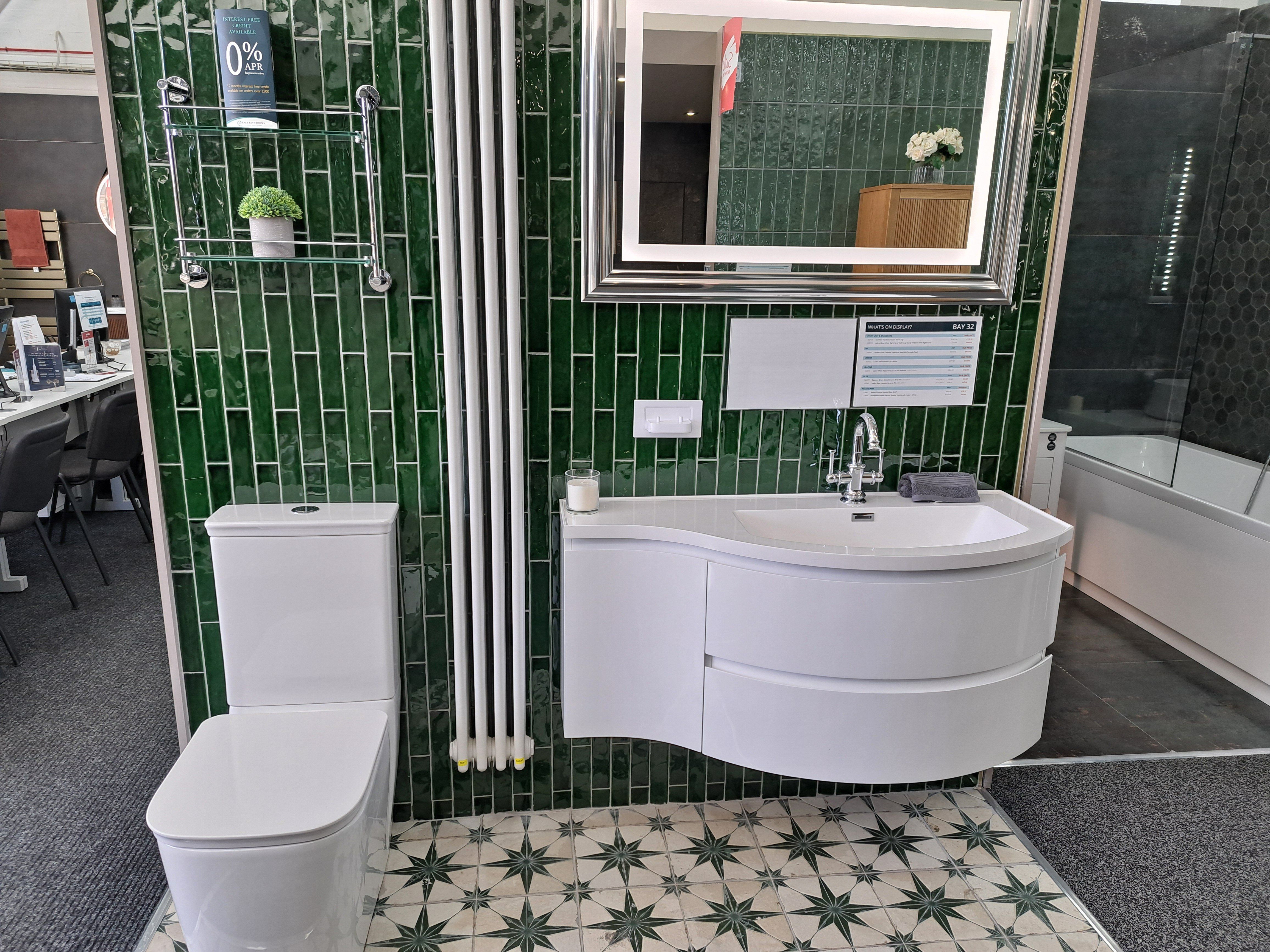 Green tiled bathroom display with a white vanity unit, toilet and illuminated mirror.