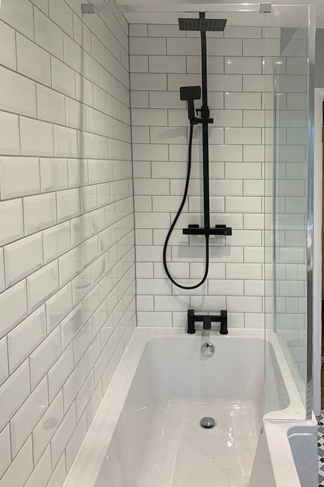 A crisp monochrome bathroom featuring white bevelled metro wall tiles and a straight bath. The setup includes a modern matt black square rainfall shower with a matching handheld head and black deck-mounted bath filler taps.