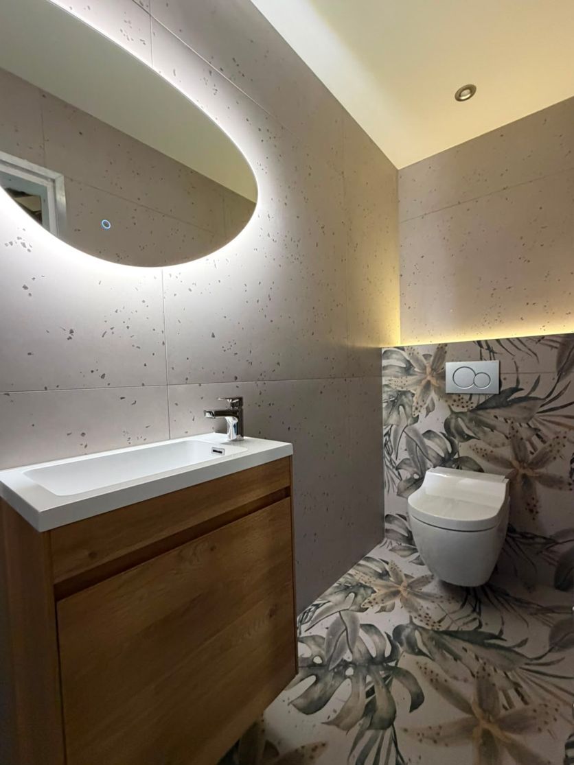 Modern bathroom featuring an Odion natural oak vanity with an Elora brushed nickel tap, an Ivy pebble LED mirror, and Dante Blanco terrazzo and tropical decor wall tiles illuminated by Smart Surface LED strip lighting.
