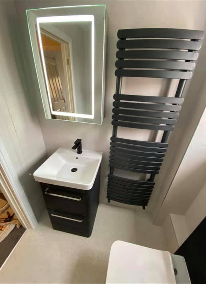 A compact modern cloakroom featuring a slimline matt black floor-standing vanity unit with a white ceramic basin and matt black tap, paired with a large curved matt black heated towel radiator and a portrait LED illuminated mirror cabinet.