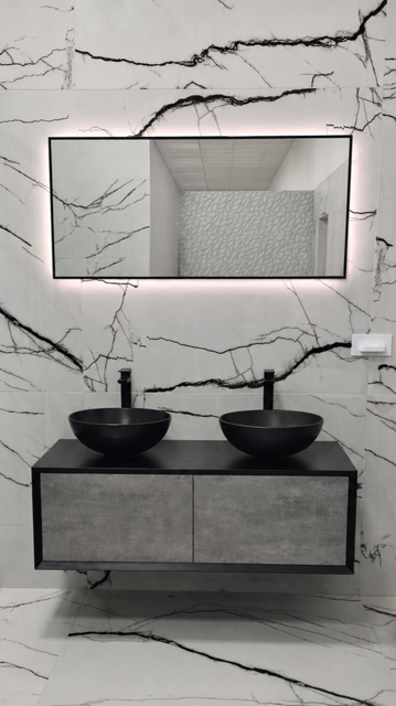 Modern double vanity display with two black vessel basins, a wall-hung concrete-effect unit, illuminated mirror and white marble-effect tiles.
