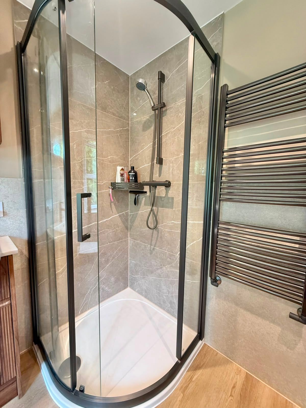 Completed bathroom shower enclosure with gunmetal frame, rainfall shower and wood-effect flooring.