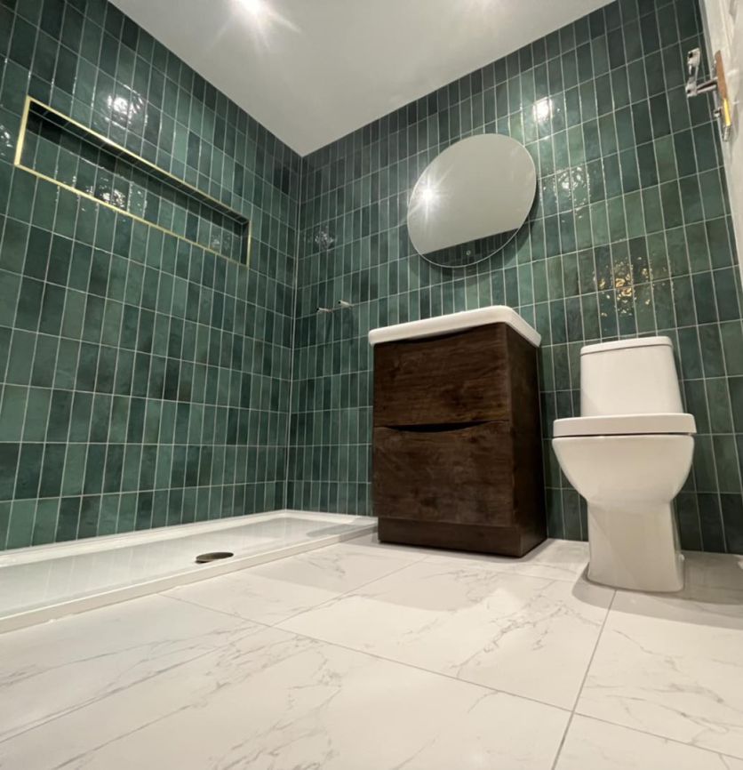 A luxurious bathroom featuring vertical-stacked emerald green glossy wall tiles with a gold-trimmed recessed niche. The space includes a dark wood floorstanding vanity unit, a modern white toilet, and large-format white marble effect floor tiles.