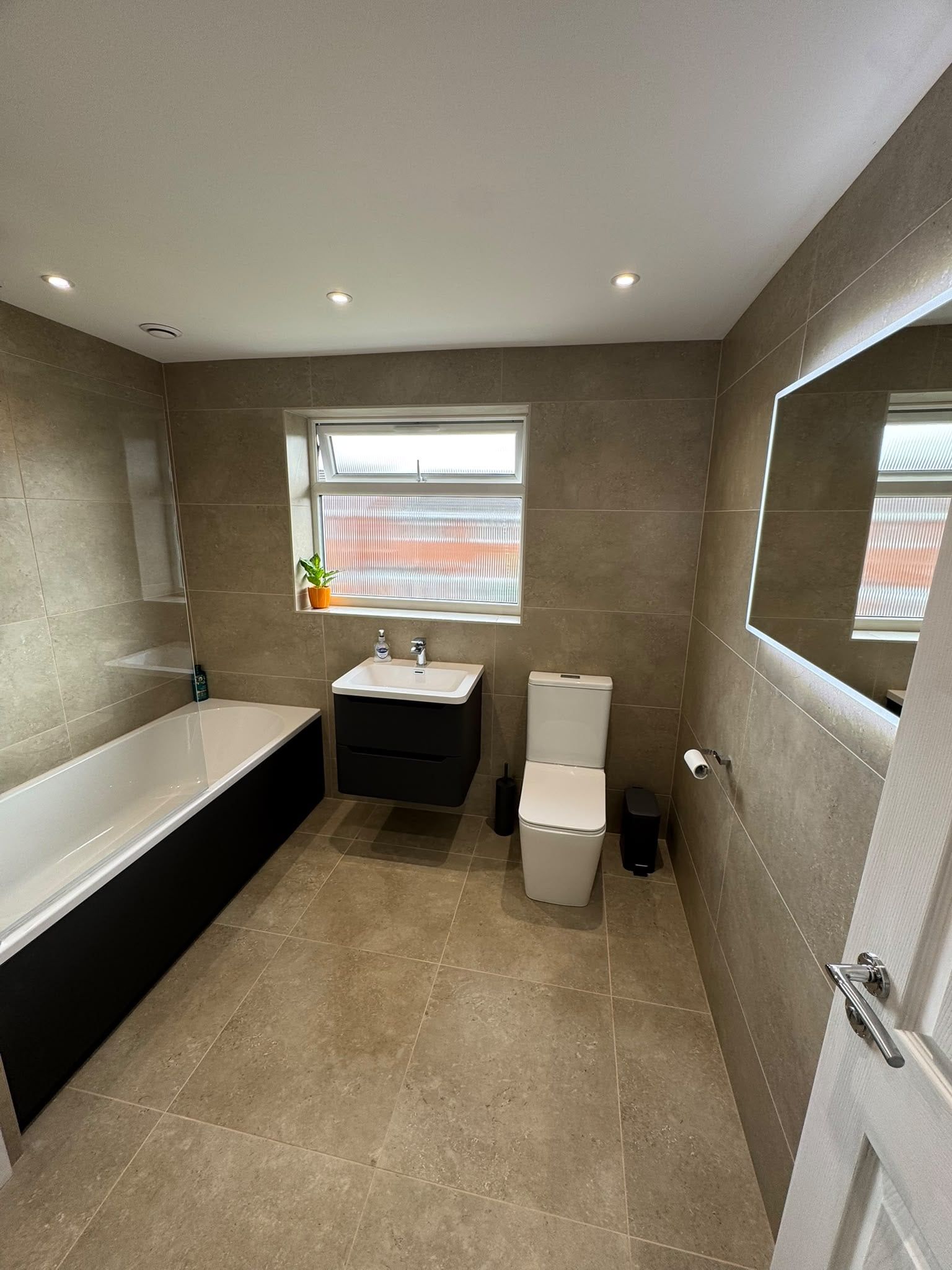 Completed modern bathroom with a matt black vanity unit, toilet, bath and warm neutral stone-effect tiles.